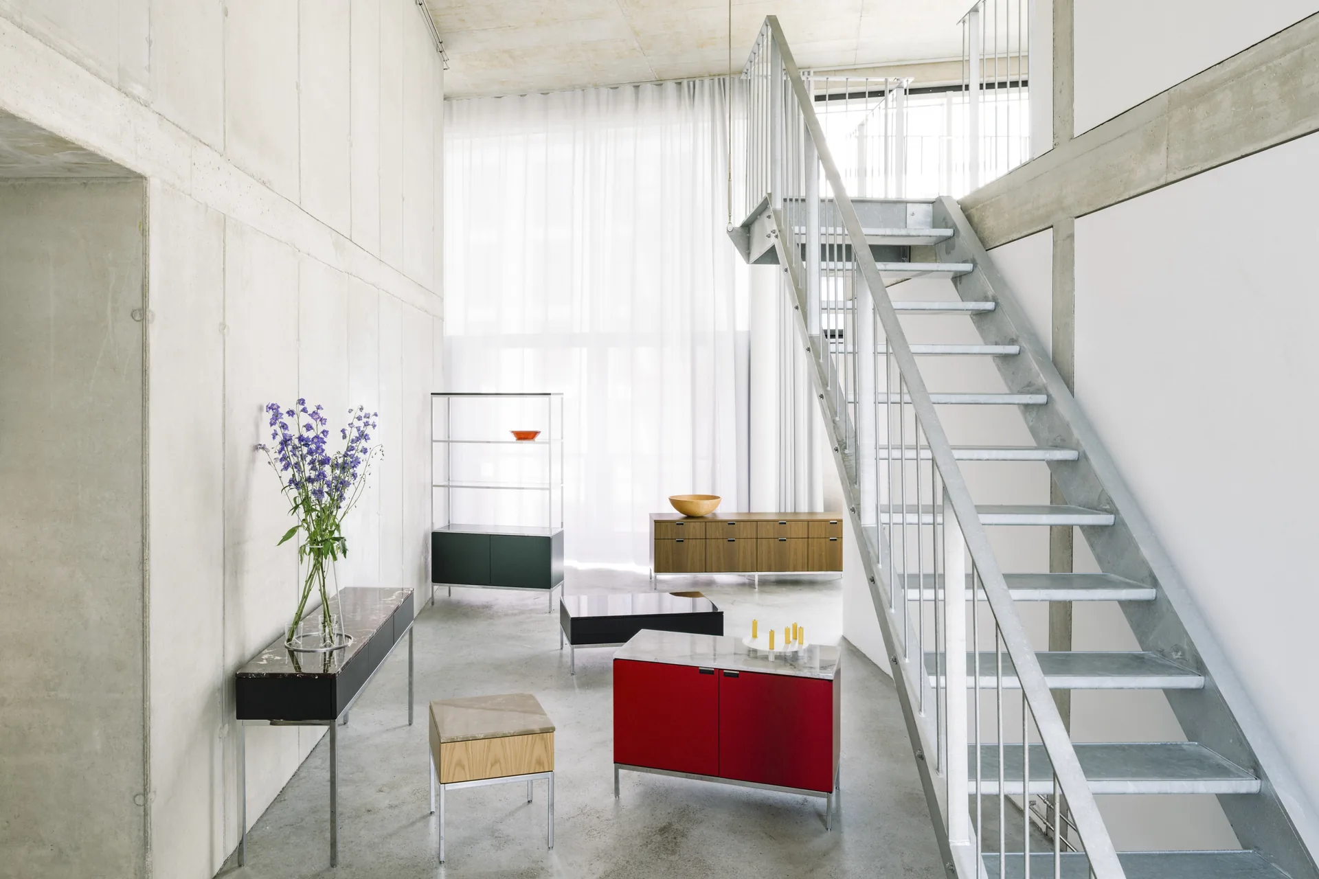 Knoll Chermayeff Storage: aparador rojo, consola negra y estantería abierta en un loft de hormigón con escalera de acero